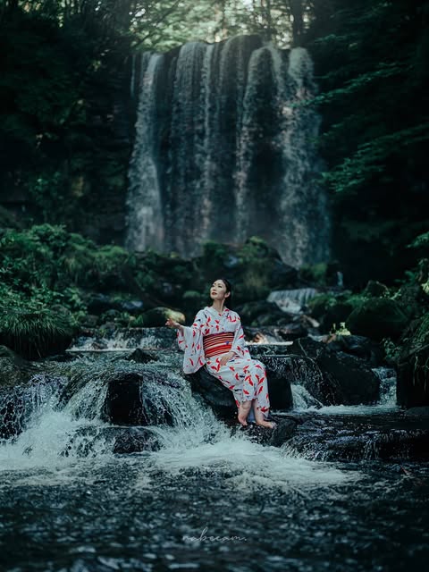 緑に包まれた滝の前で、岩に座る浴衣姿の人物。